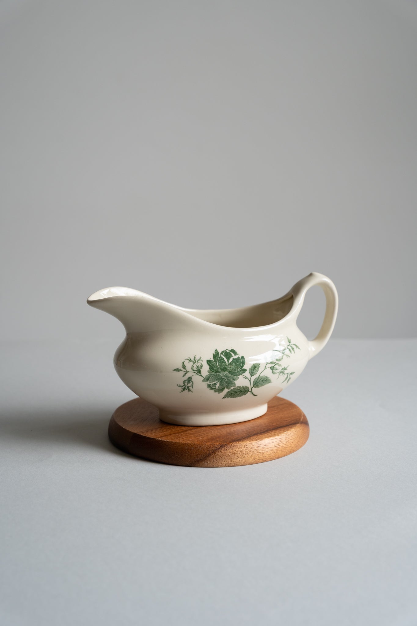White ceramic gravy boat with green floral design on a wooden stand against a gray background