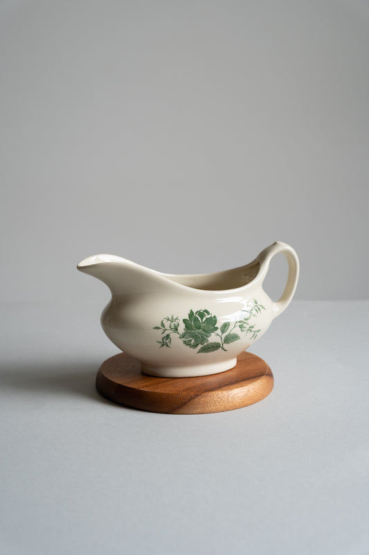 White ceramic gravy boat with green floral design on a wooden stand against a gray background